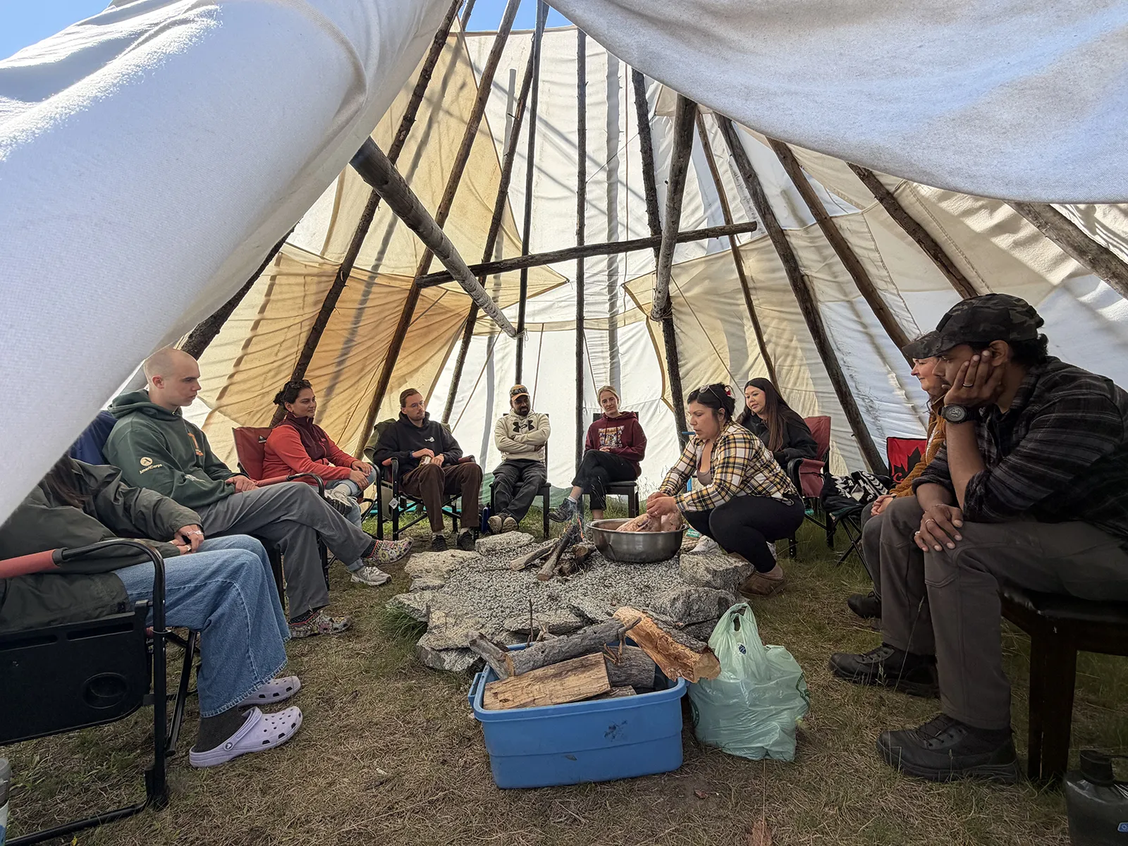 Group of people sitting in a circle inside a teepee around a fire pit, one person kneading dough.