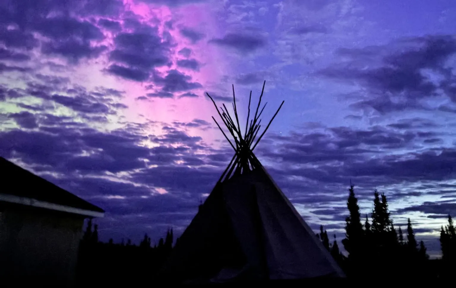 Silhouette of a tipi and trees against a vibrant purple and blue cloudy sunset sky.