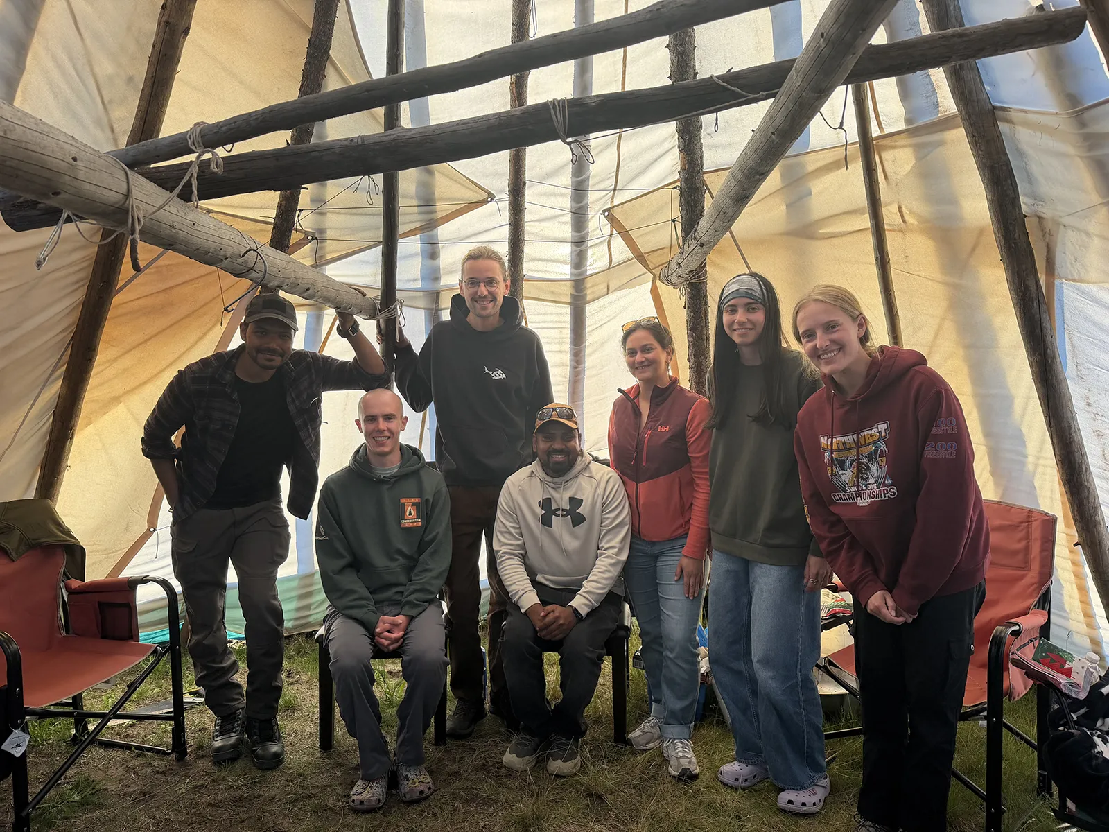 Group of seven smiling people posing inside a large tent with wooden poles and fabric walls.