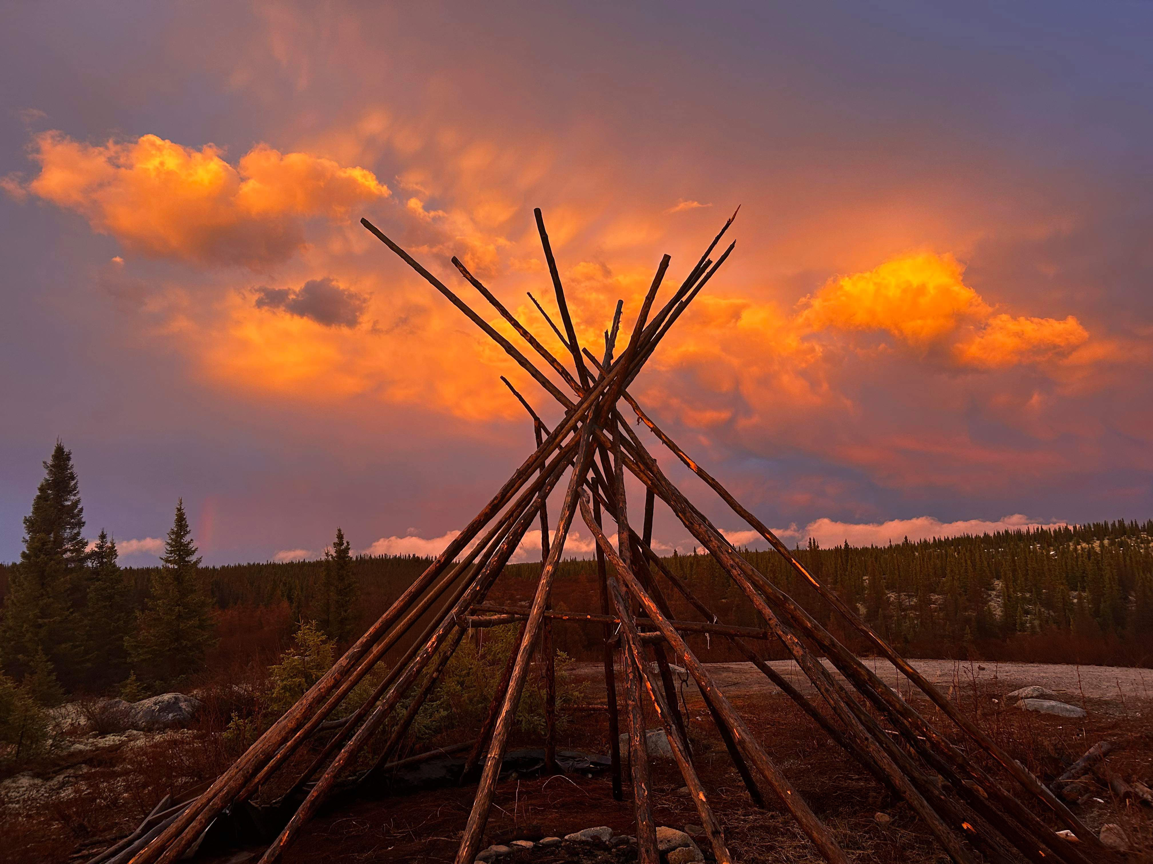 Wooden teepee frame under a glowing orange sunset sky above a forest landscape.