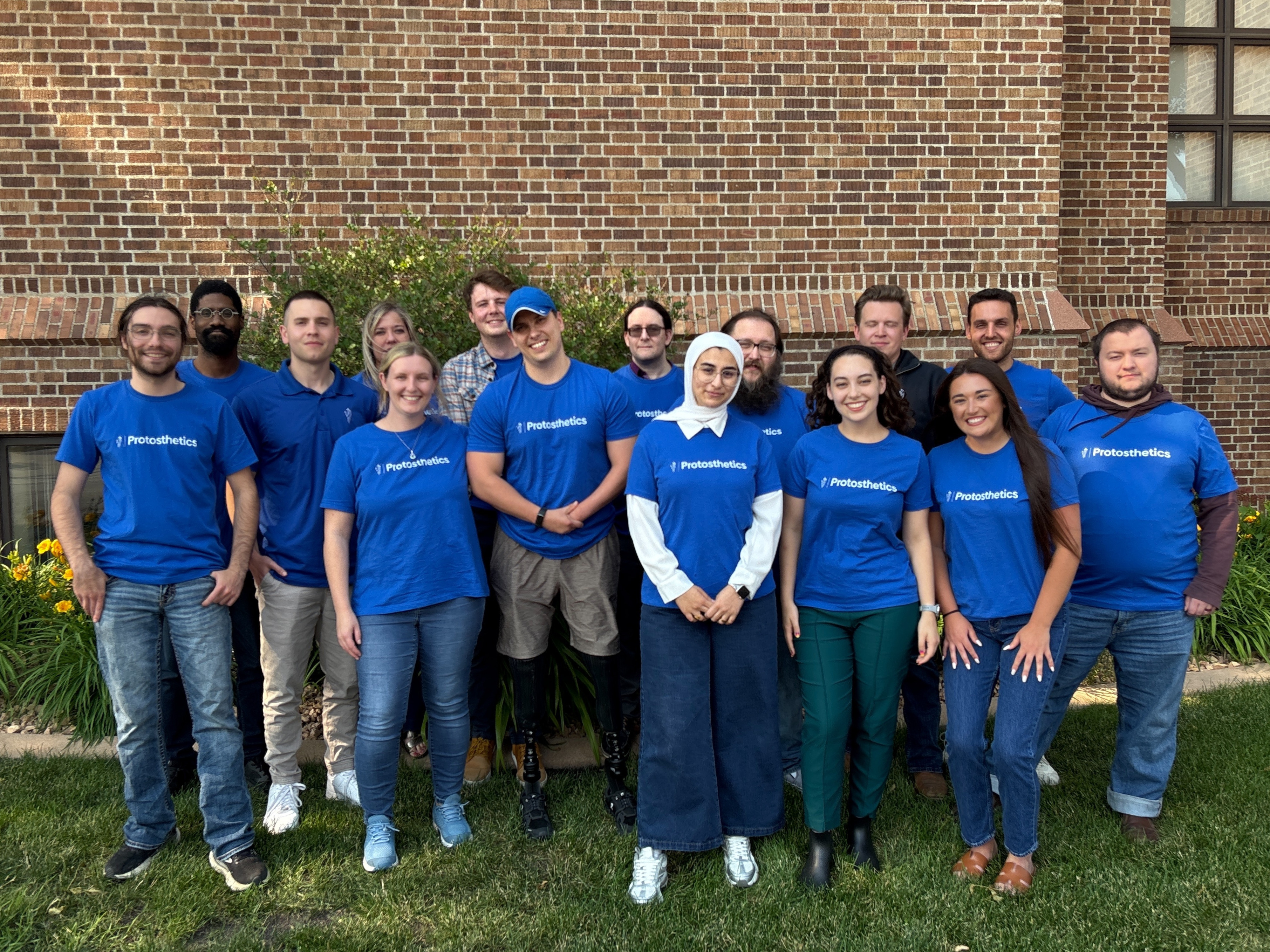 Group photo of 15 people wearing blue Protosthetics t-shirts standing outdoors in front of a brick wall.