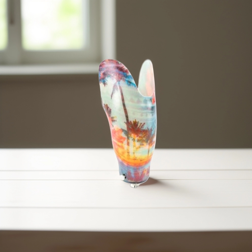 Colorful prosthetic leg socket with palm tree and sunset design on white table near window.