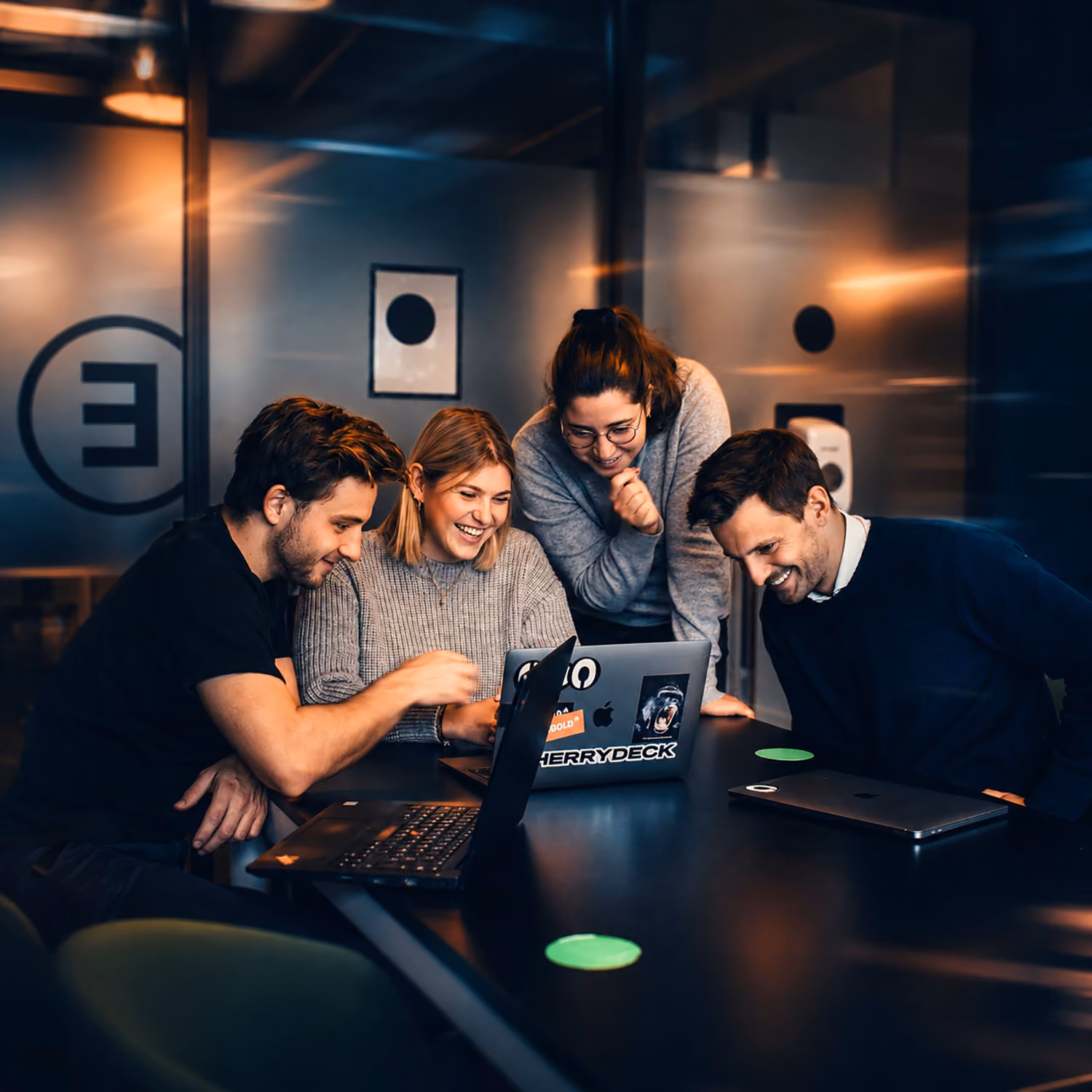 Four colleagues smiling and working together on laptops in a modern office meeting room.