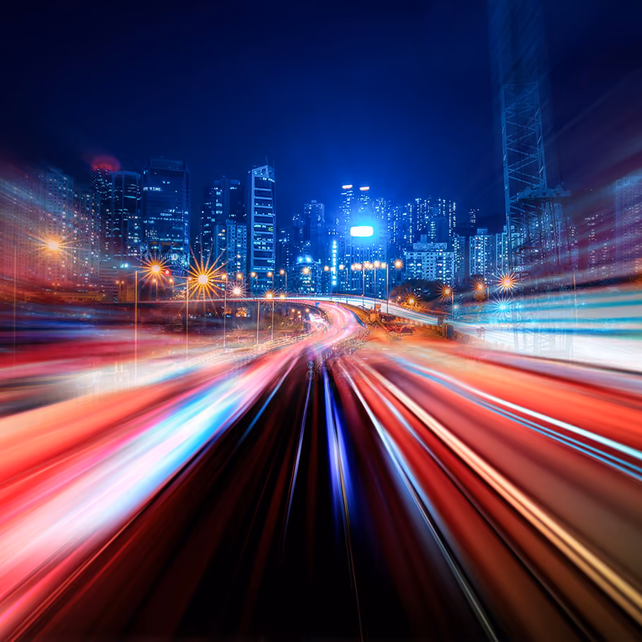 Blurred light trails on a highway at night with a lit city skyline in the background.