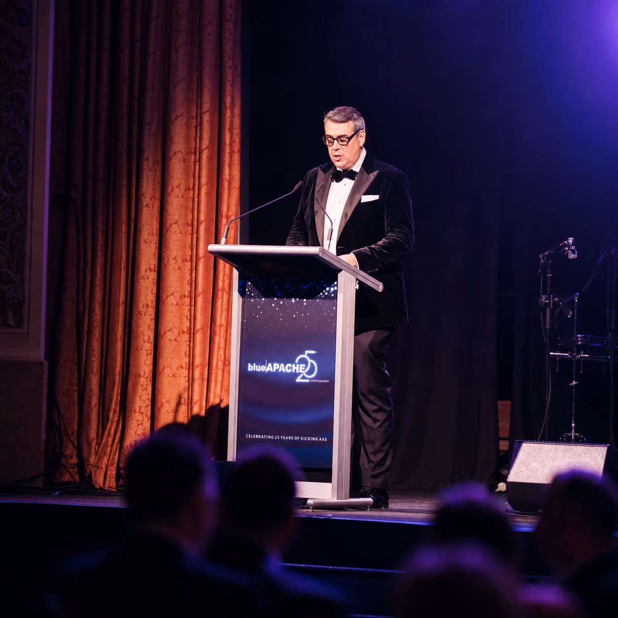 Man in tuxedo speaking at a podium with blueAPACHE 25th anniversary logo on stage.