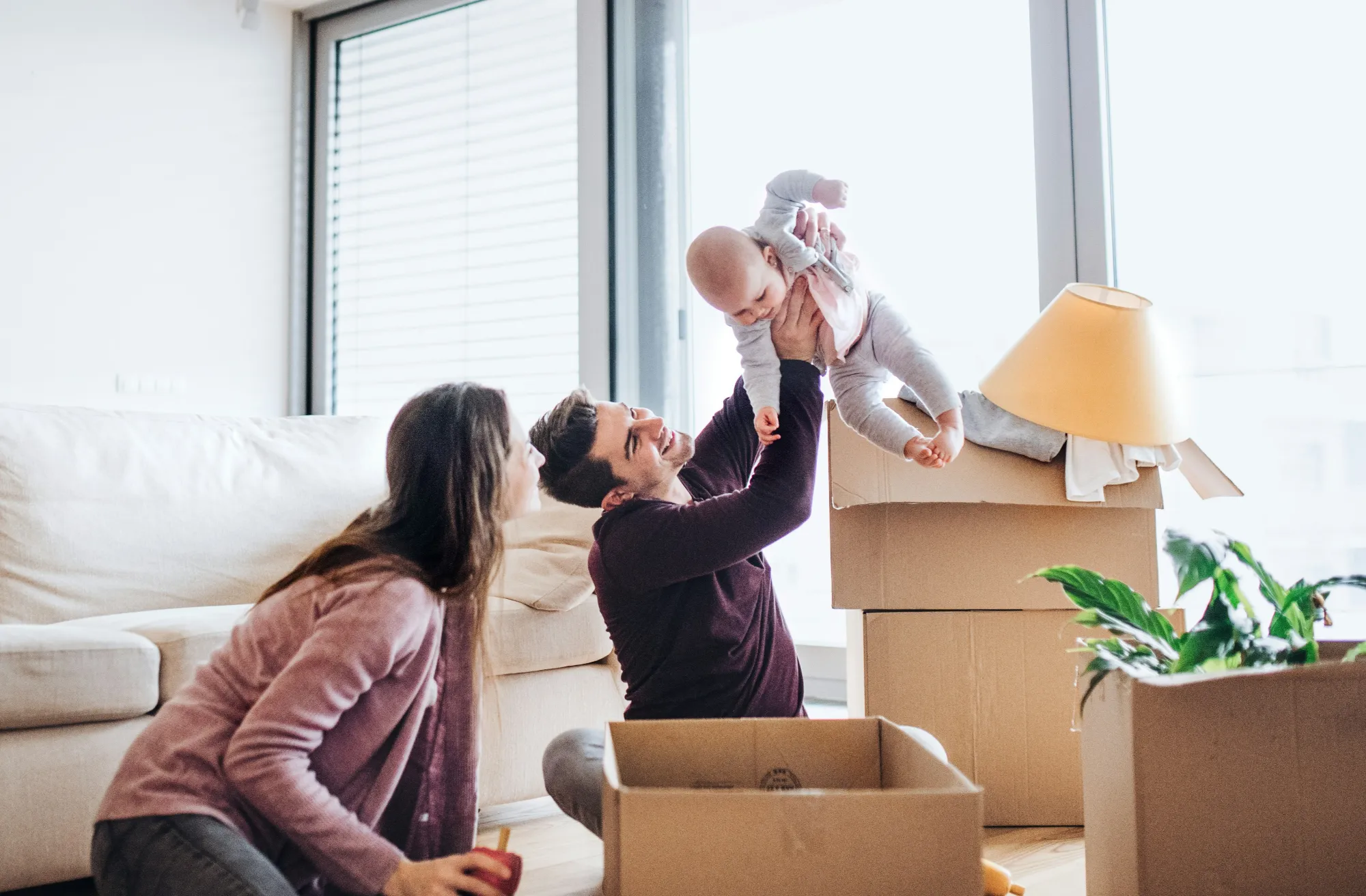 Man holding baby over cardboard boxes while woman watches in a bright living room.