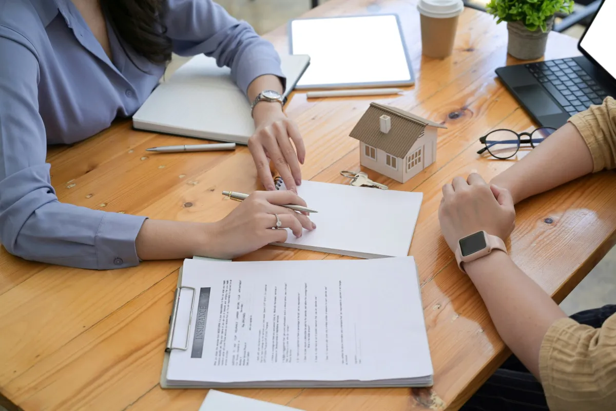 Two people signing document at table with house model, keys, laptop, and coffee cup nearby.
