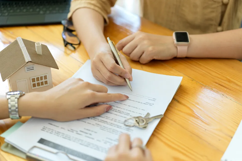 Two people signing a real estate contract with a house model and keys on a wooden table.