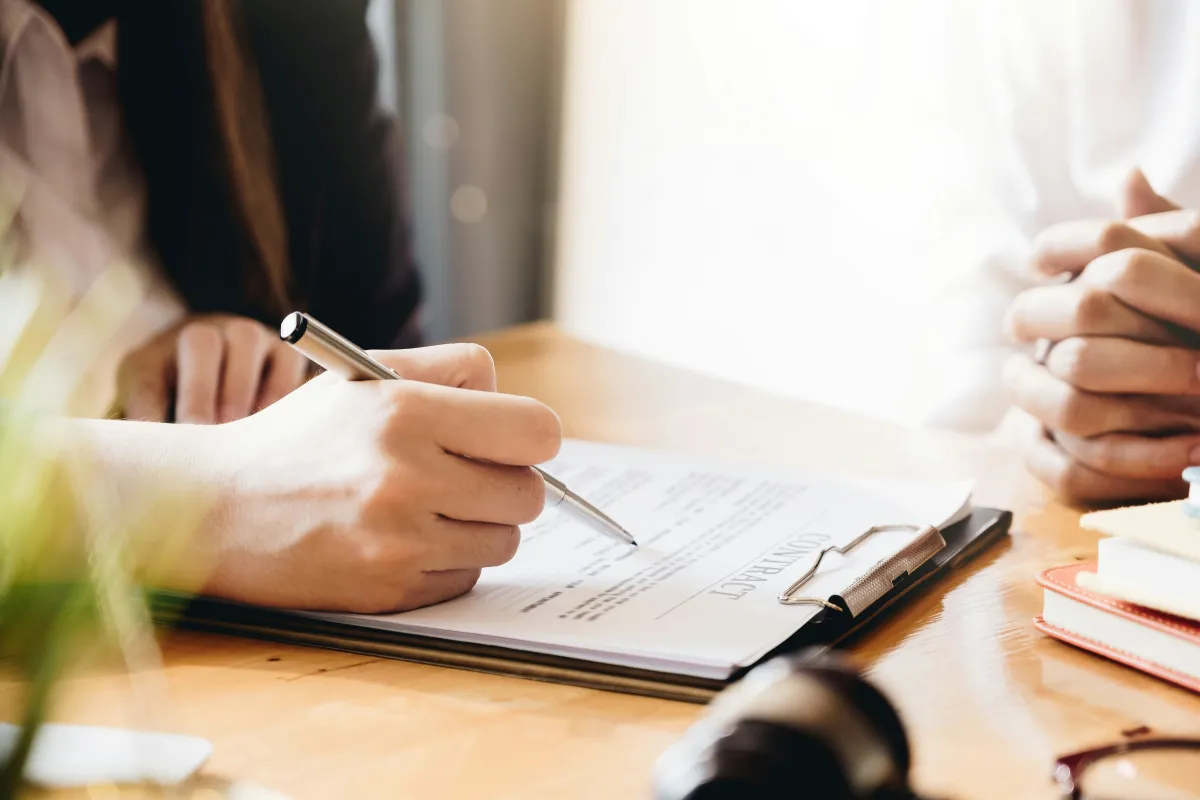 Person signing a contract document on a clipboard at a wooden desk with another person's hands visible.
