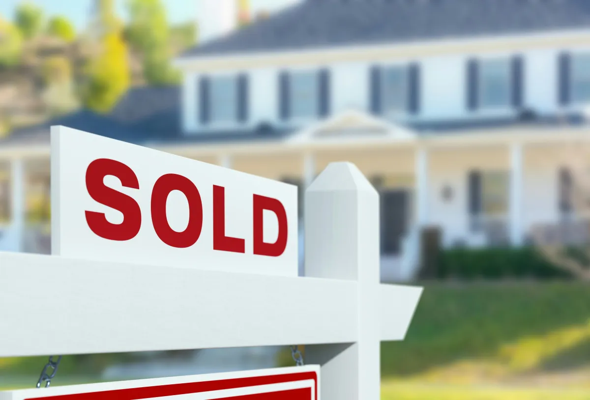 White sold sign in front of a large house with green lawn and trees in the background.