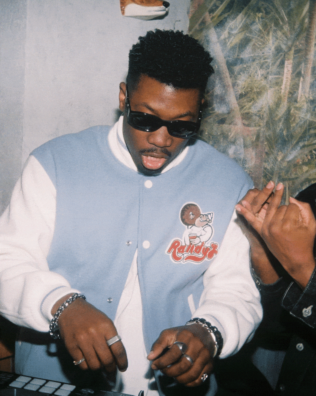 Man wearing sunglasses and a blue and white jacket with Randy's logo using DJ equipment.