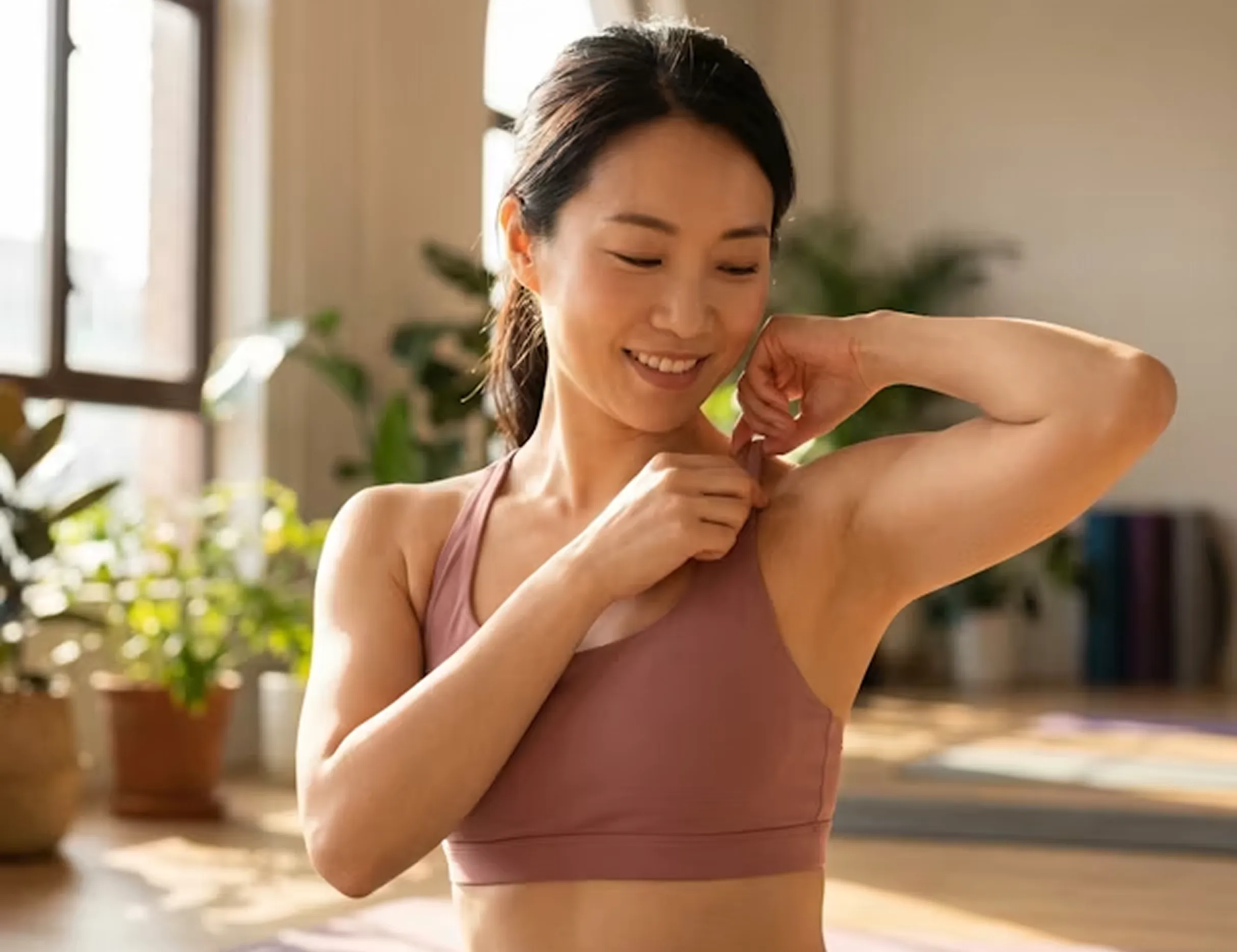 Smiling woman in workout top adjusting strap in sunlit room with plants and yoga mats.