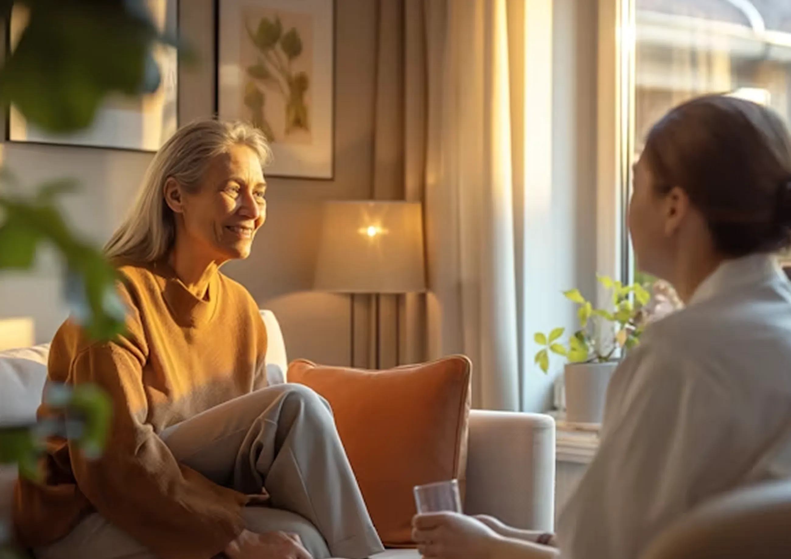 Two women sitting and talking warmly on a couch in a sunny, cozy living room.