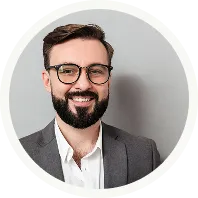 Smiling man with beard and glasses wearing a gray blazer and white shirt against a light background.