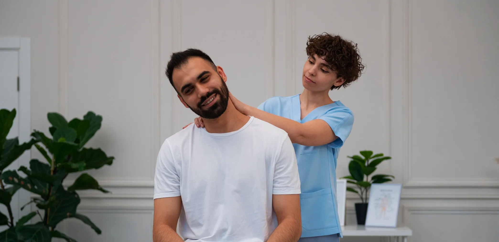 Physiotherapist in blue scrubs stretching a man's neck in a bright clinic room with plants.