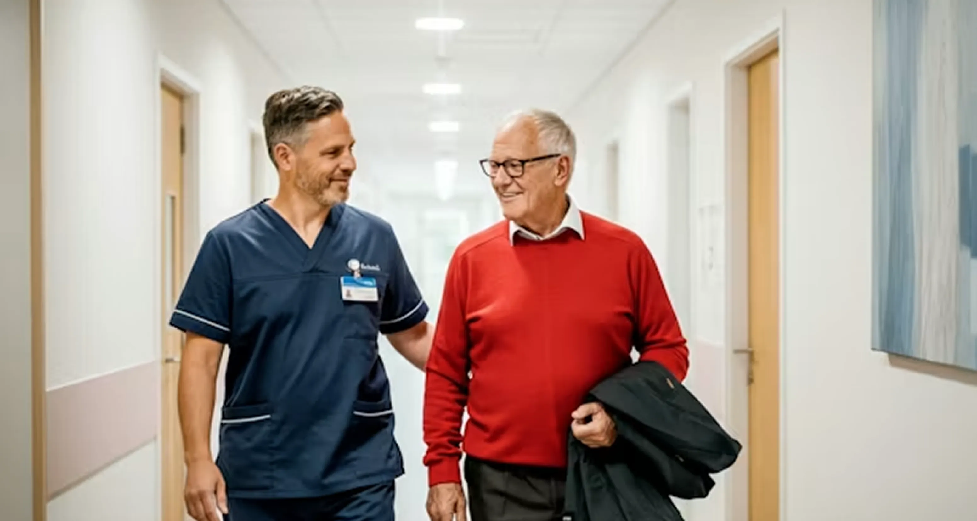 Nurse in navy scrubs walking and smiling with elderly man in red sweater in hospital hallway.