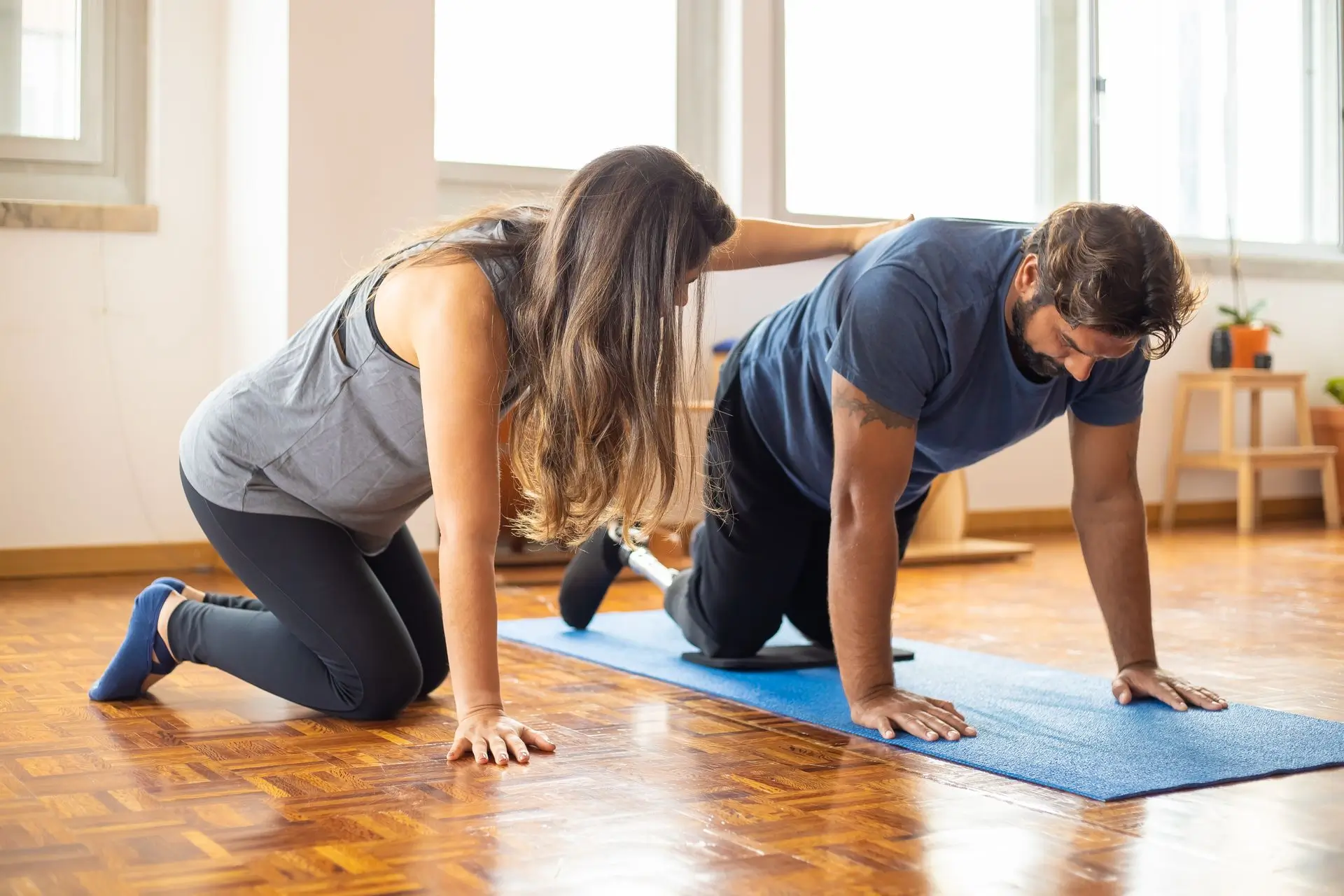 Woman assisting man with prosthetic leg doing an exercise on a blue mat indoors.