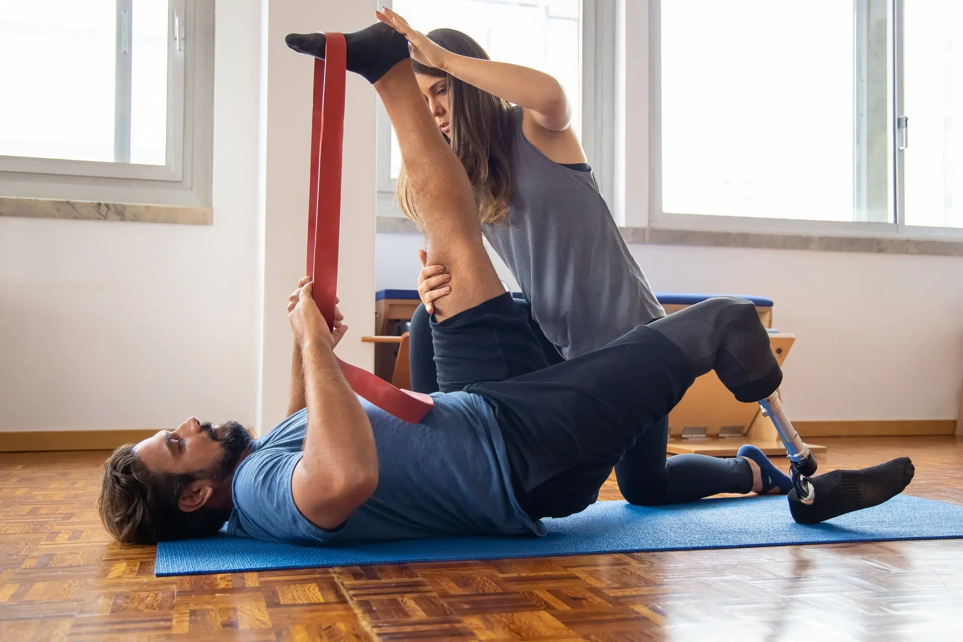 Therapist assists man with prosthetic leg doing leg stretch using resistance band on mat.