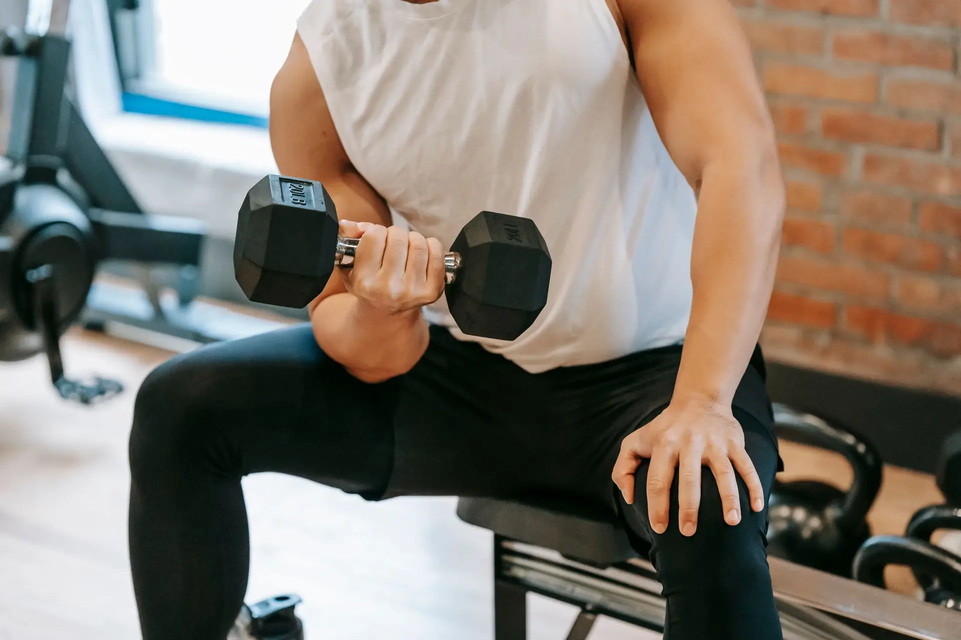 Man wearing white sleeveless shirt lifting a 20-pound dumbbell while seated in a gym.