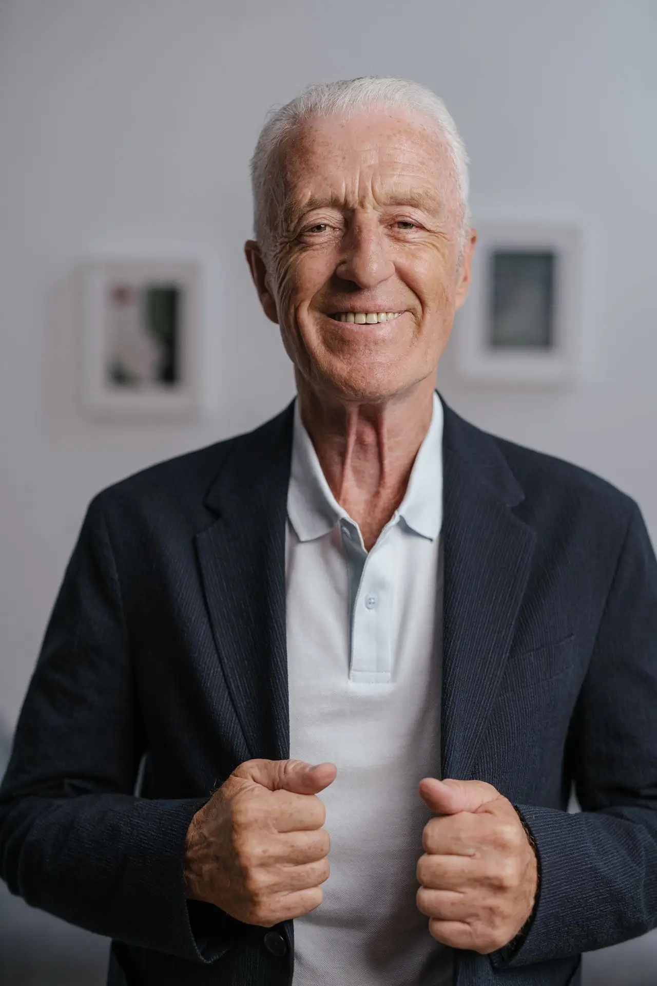Smiling elderly man wearing a navy blazer and white polo shirt standing indoors.