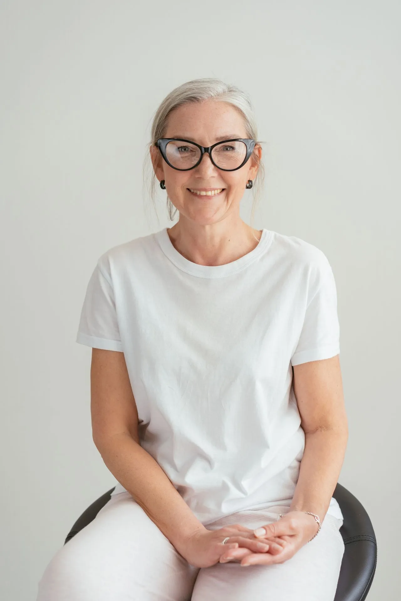 Smiling older woman with gray hair and black glasses wearing a white t-shirt sitting on a black chair.