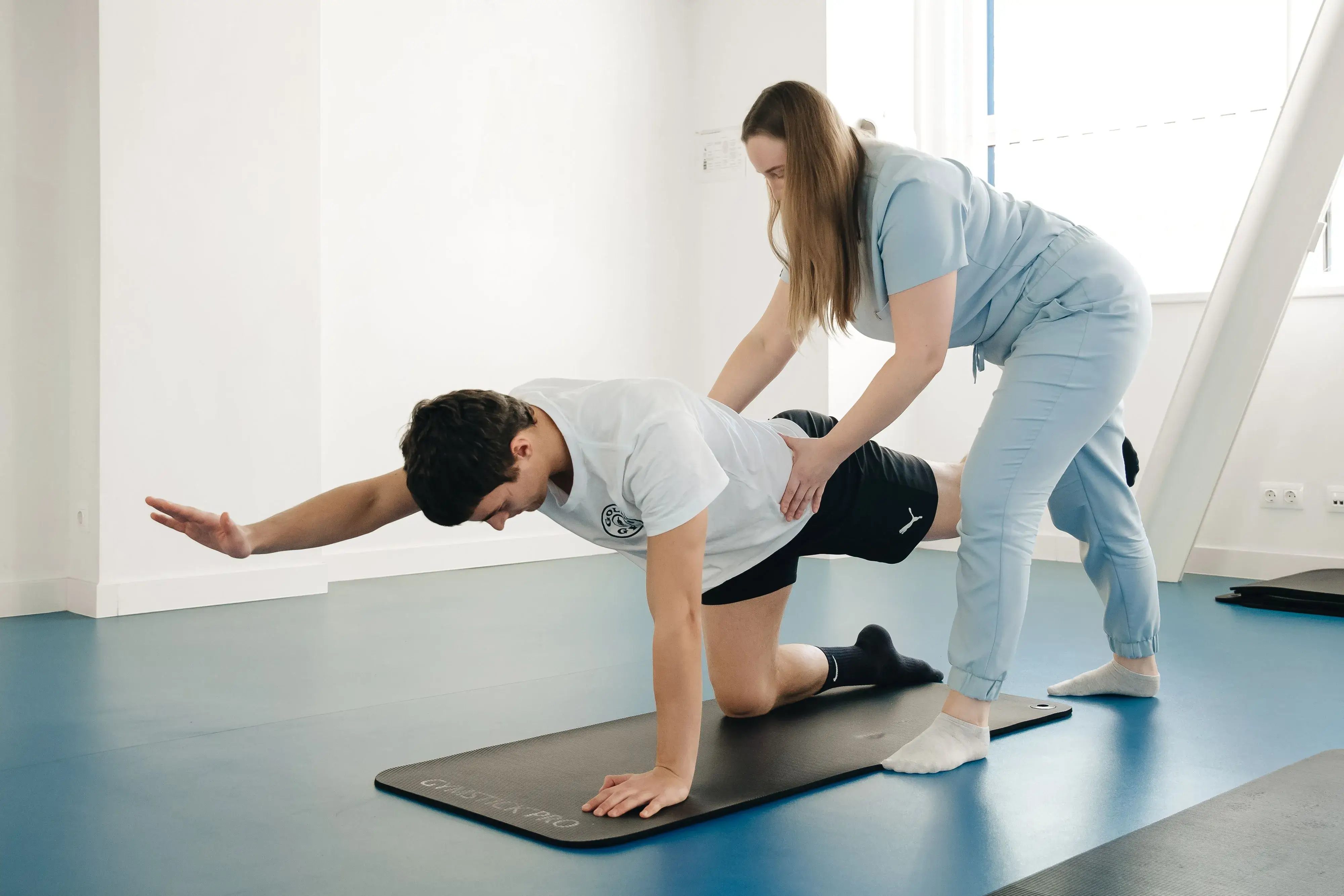 Physical therapist assists man in balancing on one hand and opposite knee on exercise mat.