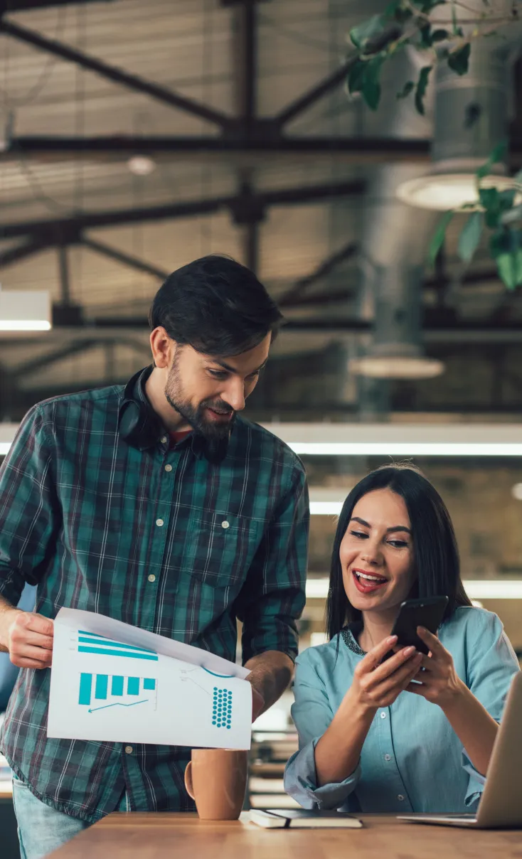 A man in a checkered shirt holding documents with charts stands next to a woman who is smiling and looking at her phone in an office setting.