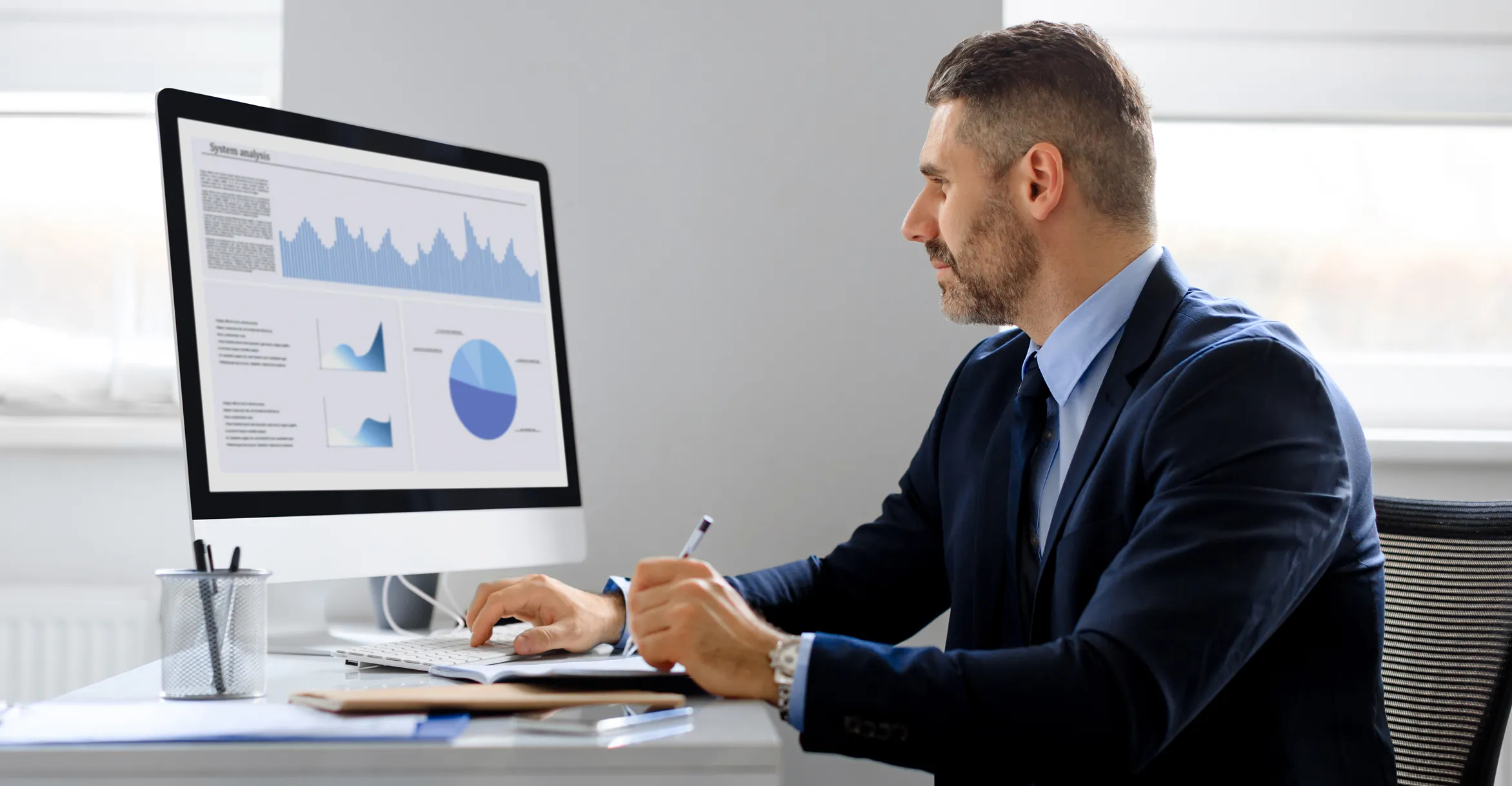 A man in a blue suit sitting at a desk, analyzing data charts displayed on a desktop computer screen, including bar graphs, line charts, and a pie chart.