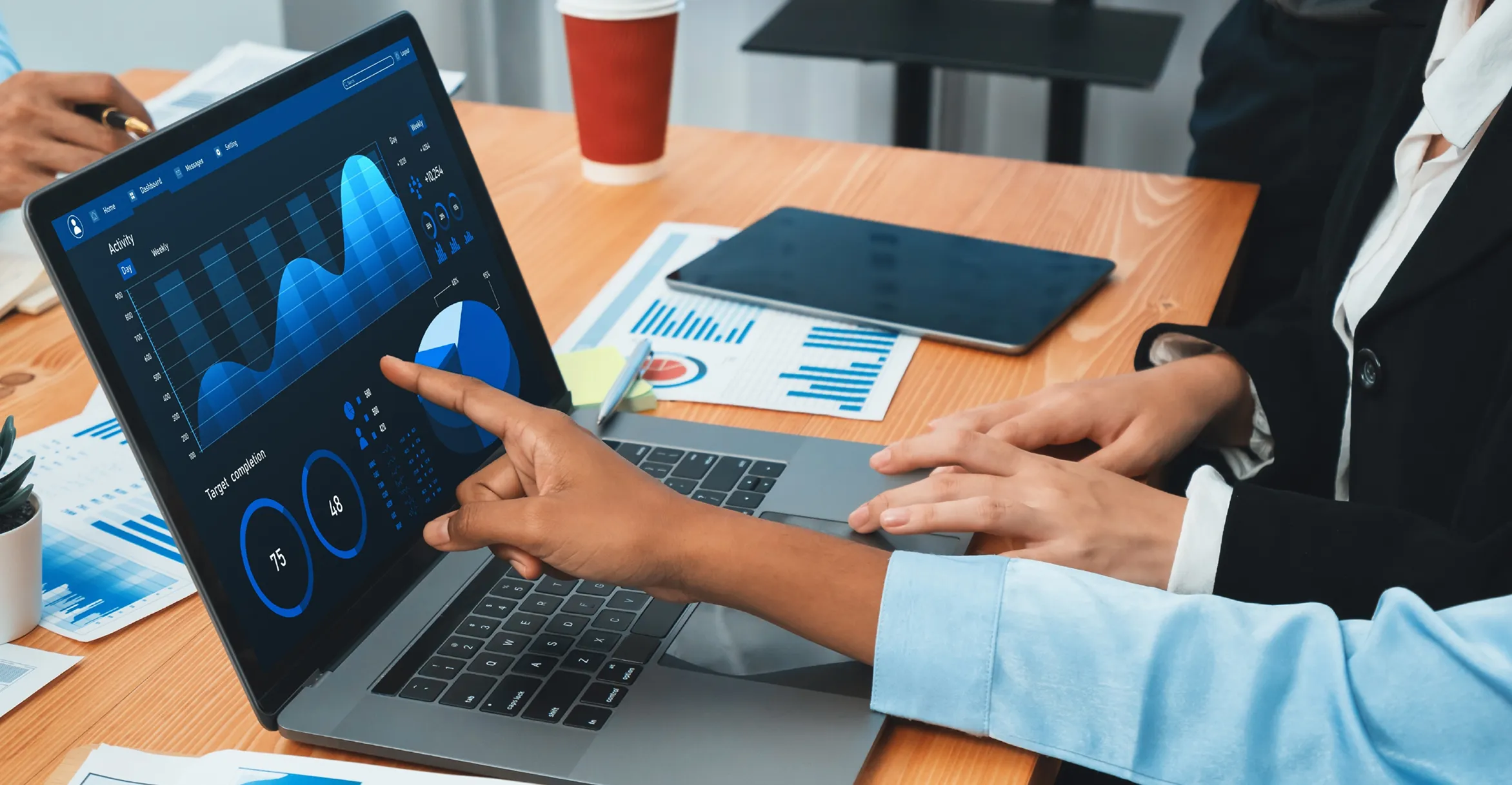 Two business professionals analyzing data on a laptop screen showing blue-themed charts and graphs including a line graph, pie chart, and circular progress indicators, on a wooden desk with a tablet, papers, and a red disposable coffee cup nearby.
