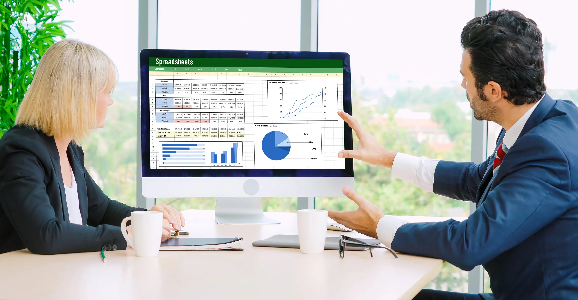 Two business professionals, a woman and a man, sitting at a table with coffee cups, looking at a large desktop screen displaying spreadsheets with tables, bar charts, pie chart, and line graph.