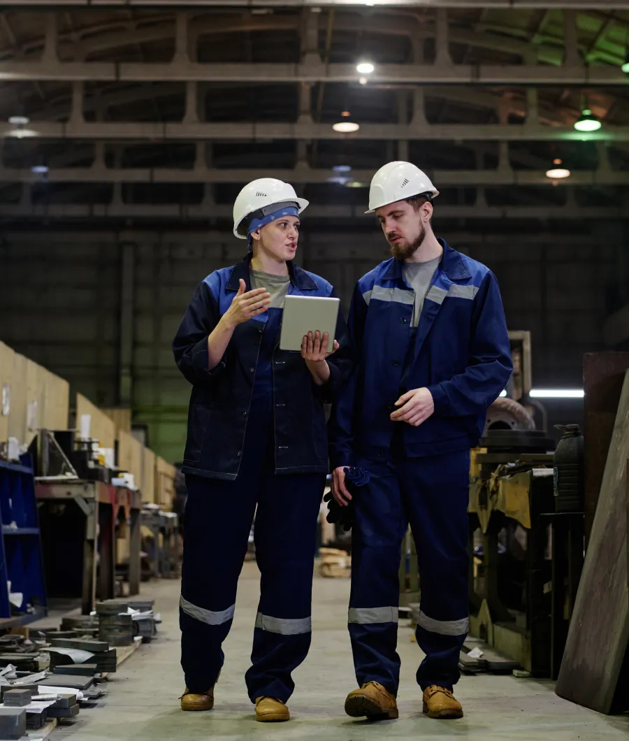 Two factory workers wearing white hard hats and navy blue uniforms walk and discuss something while looking at a tablet inside an industrial facility with machinery and equipment around.