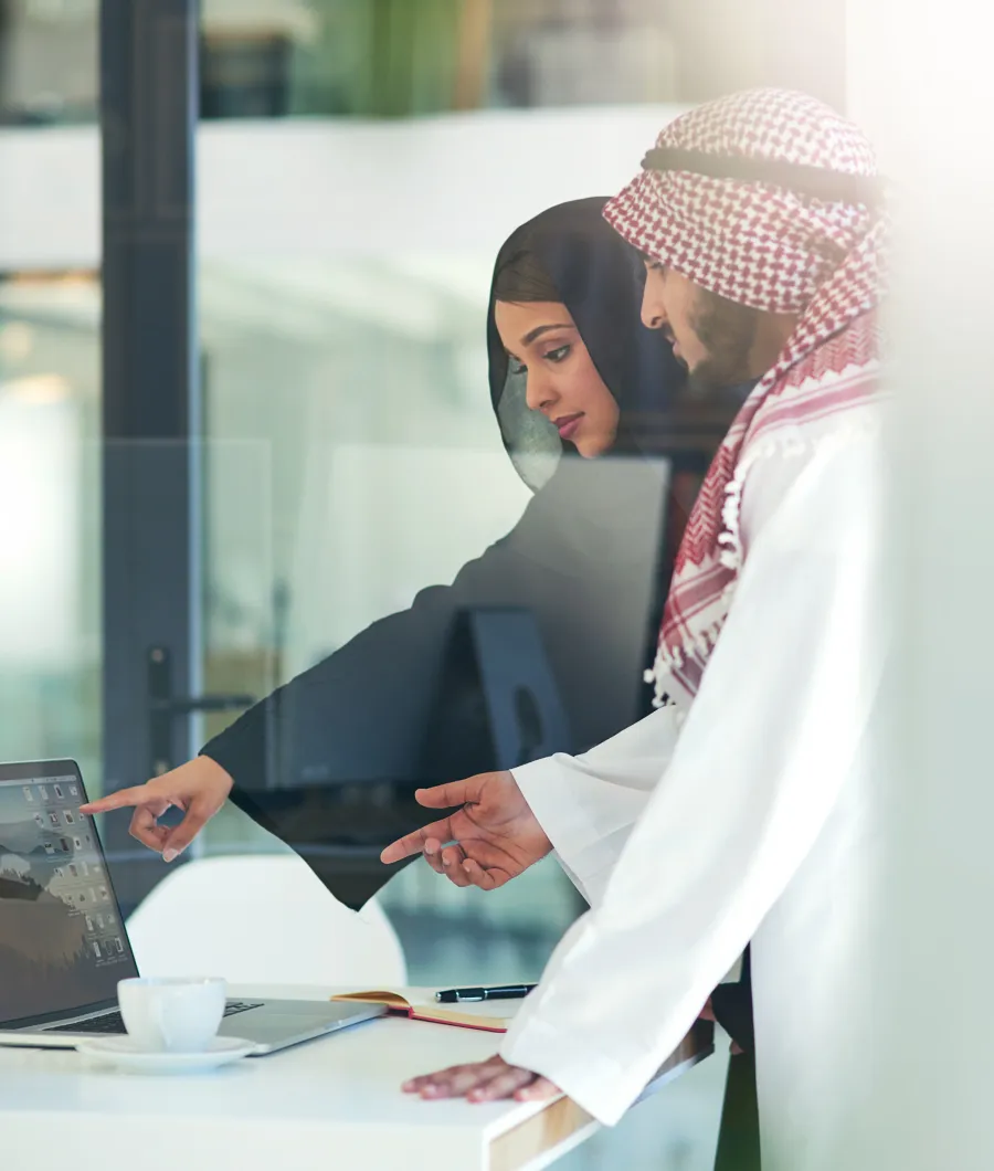 A man and a woman in traditional Middle Eastern attire looking at a laptop screen while discussing, the woman points at the screen and the man gestures with his hand.
