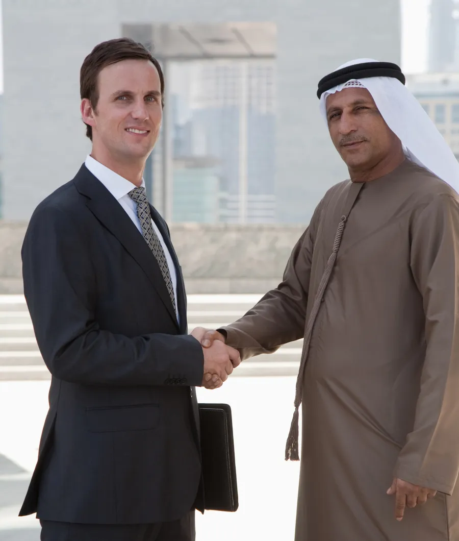 Two men shaking hands outdoors in a business setting; one man wears a dark suit and tie while holding a black folder, the other wears traditional Middle Eastern attire including a white head covering.