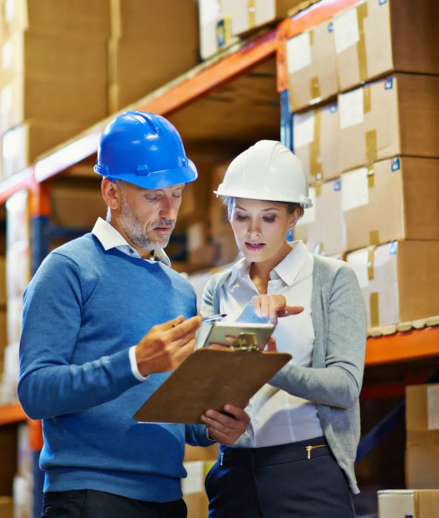 A man and woman wearing hard hats in a warehouse are reviewing a clipboard and tablet, surrounded by shelves stacked with cardboard boxes.