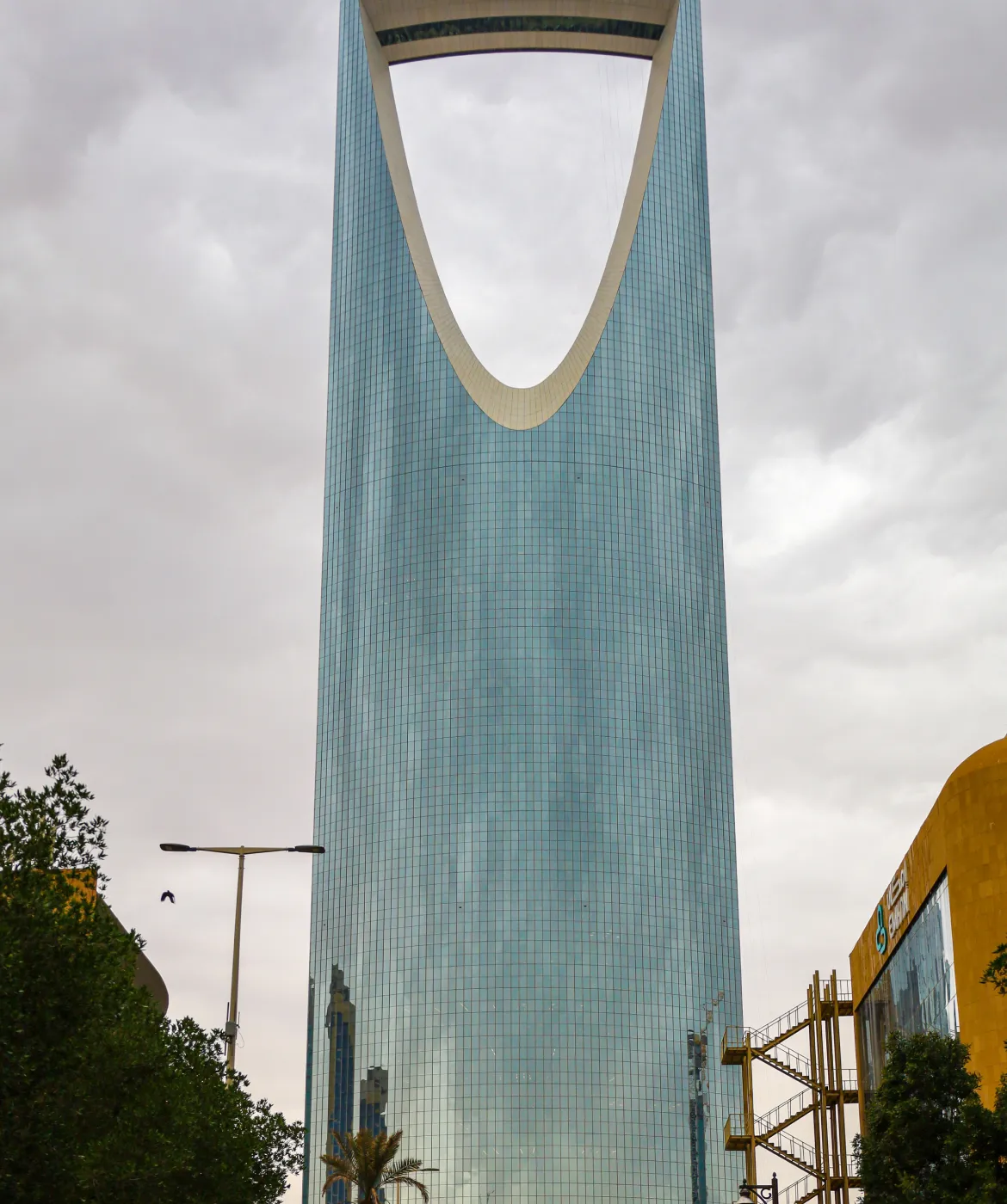 The Kingdom Centre Tower in Riyadh, Saudi Arabia, a tall modern skyscraper with a distinctive large arch near the top, reflecting the cloudy sky and nearby buildings.