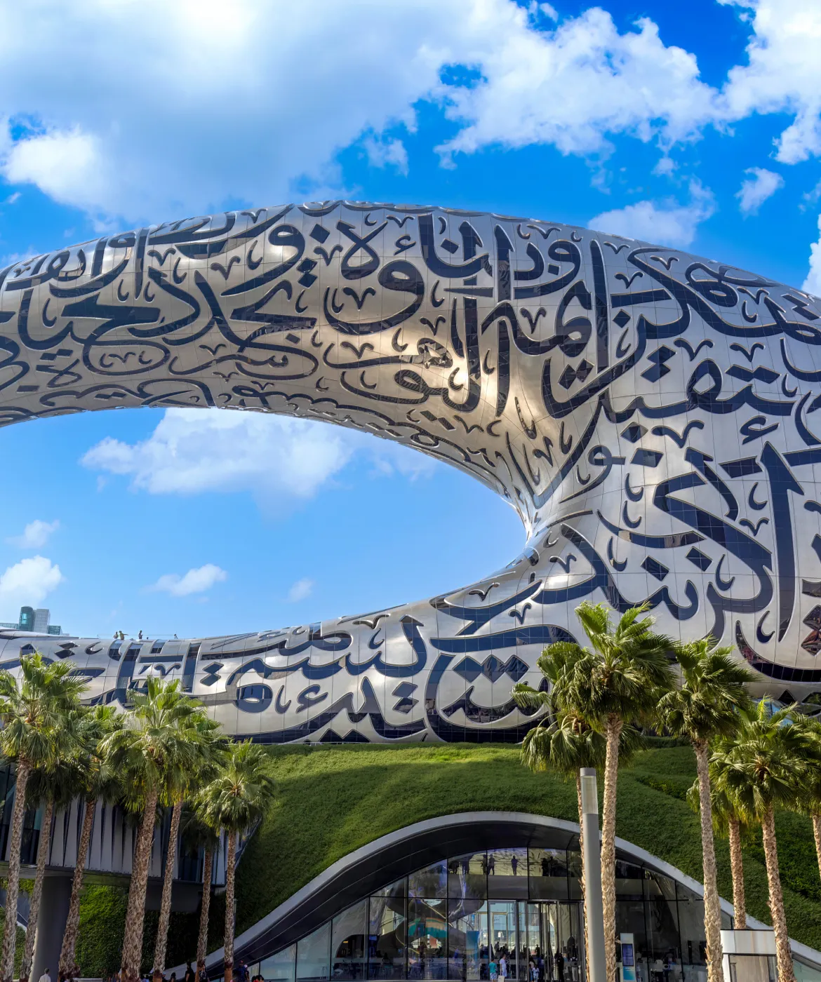 The Museum of the Future in Dubai, a futuristic torus-shaped building with Arabic calligraphy on its metallic surface, a green grassy base, palm trees, and a blue sky with clouds.