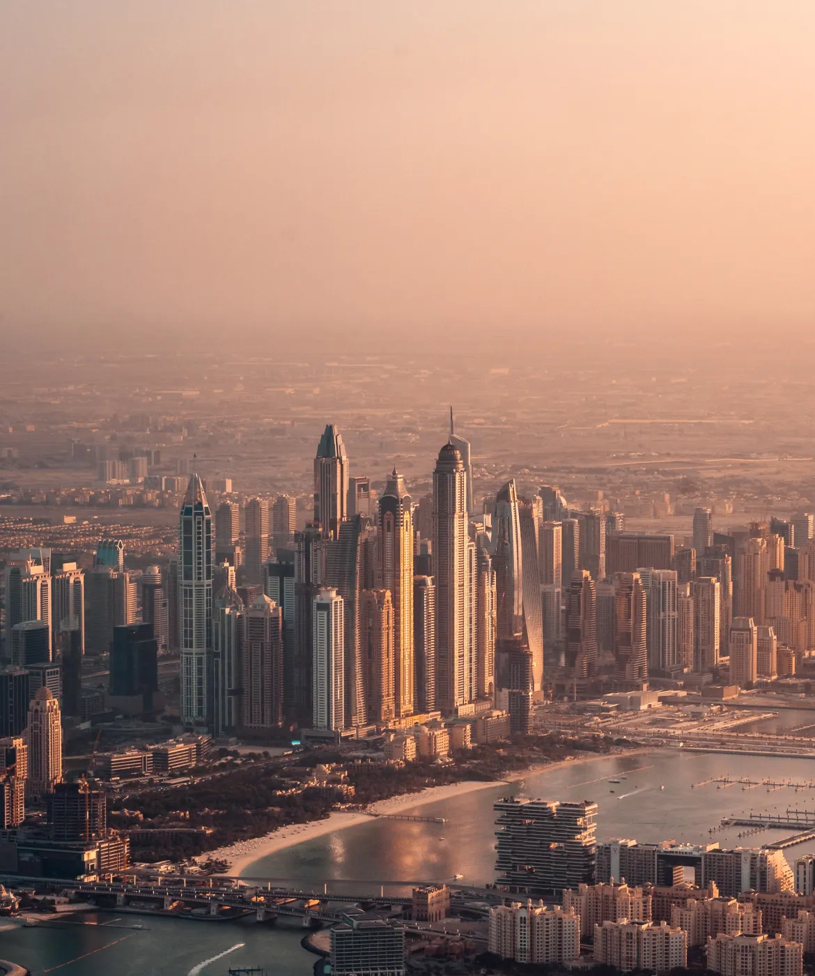 Aerial view of a modern city skyline at sunset with tall skyscrapers and a waterfront featuring a curved sandy beach and marina.