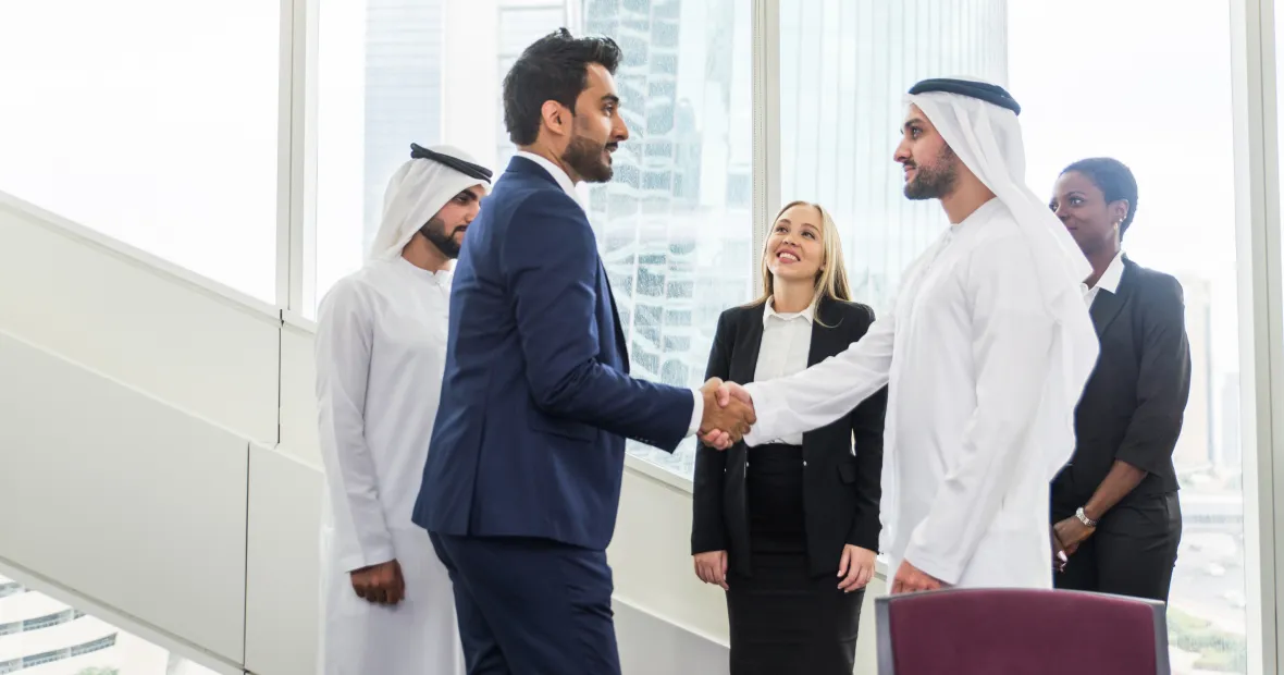 Two men, one wearing a dark blue suit and the other traditional white Middle Eastern attire, shake hands in a modern office with large windows, while three colleagues stand nearby observing and smiling.