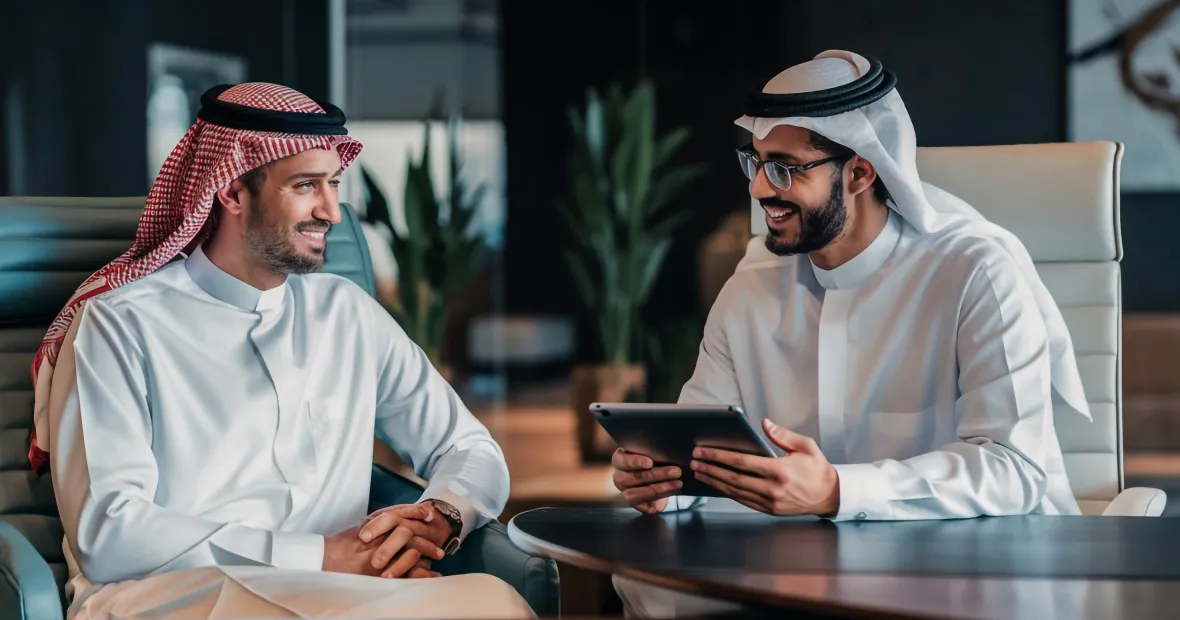 Two Arab men in traditional white clothing and head coverings sitting across from each other at a desk, one holding a tablet, both smiling in a modern office setting.