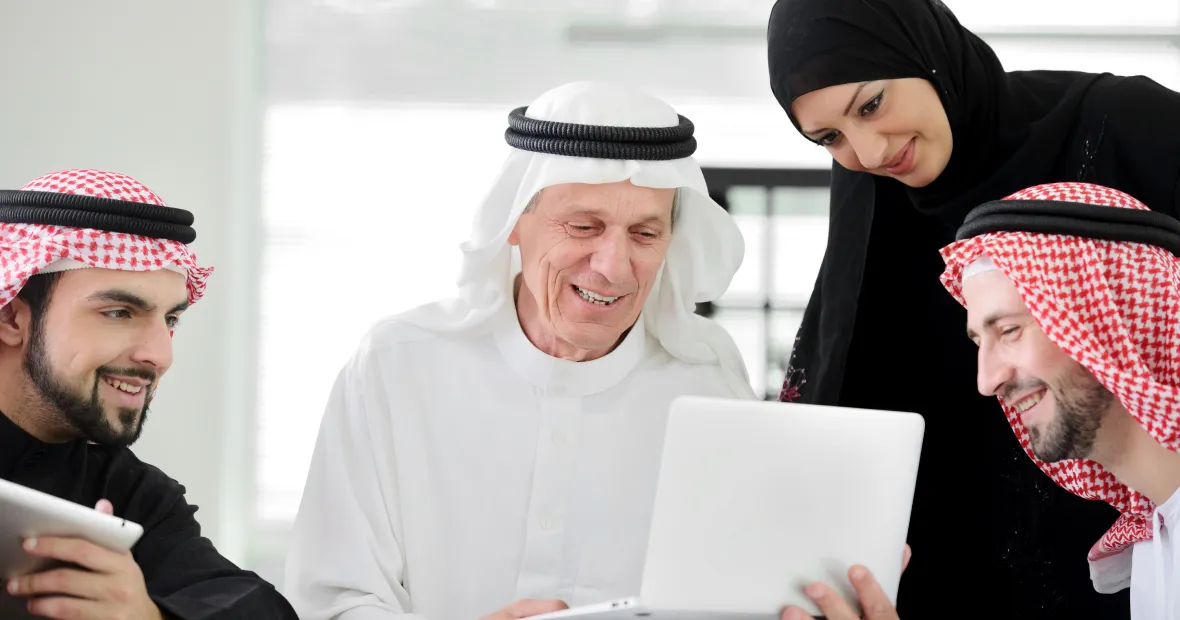 Four Arab adults gathered around a laptop with smiles, appearing engaged and collaborative in a bright indoor setting.