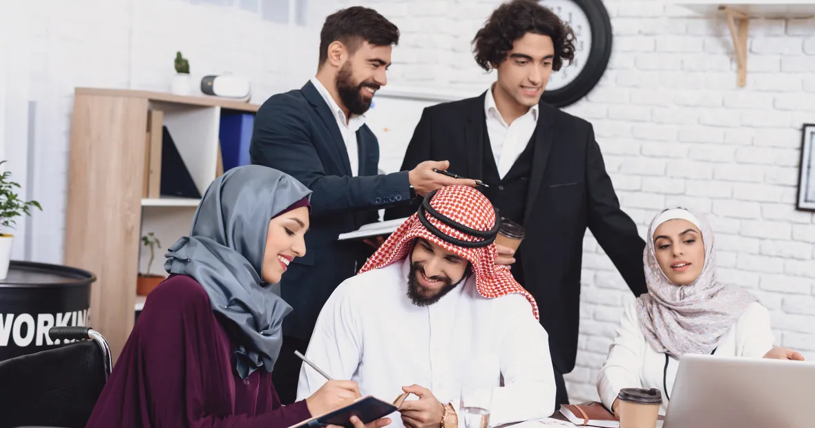 Group of five diverse professionals collaborating in a modern office, two women in hijabs working on tablets and laptops while three men in formal attire discuss and share ideas.