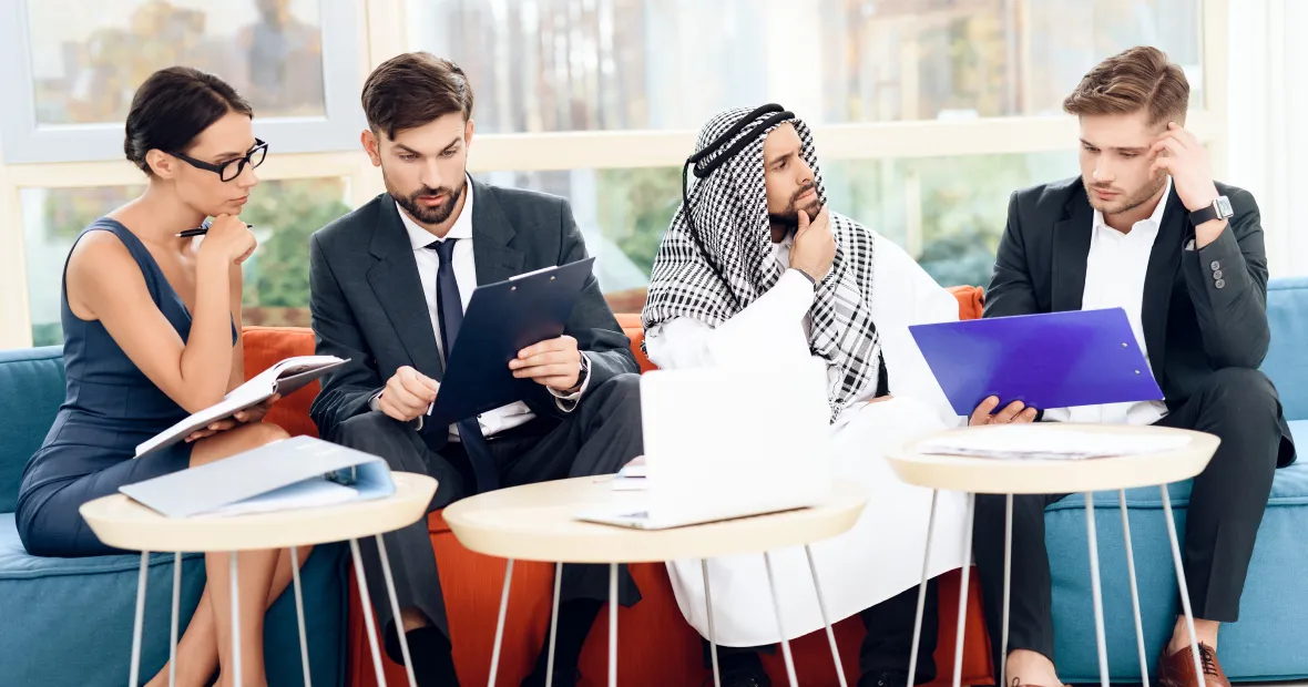 Four business professionals sitting on a couch in an office, reviewing documents; woman on left in glasses holding notes, two men in suits with clipboards, and one man in traditional Middle Eastern attire.