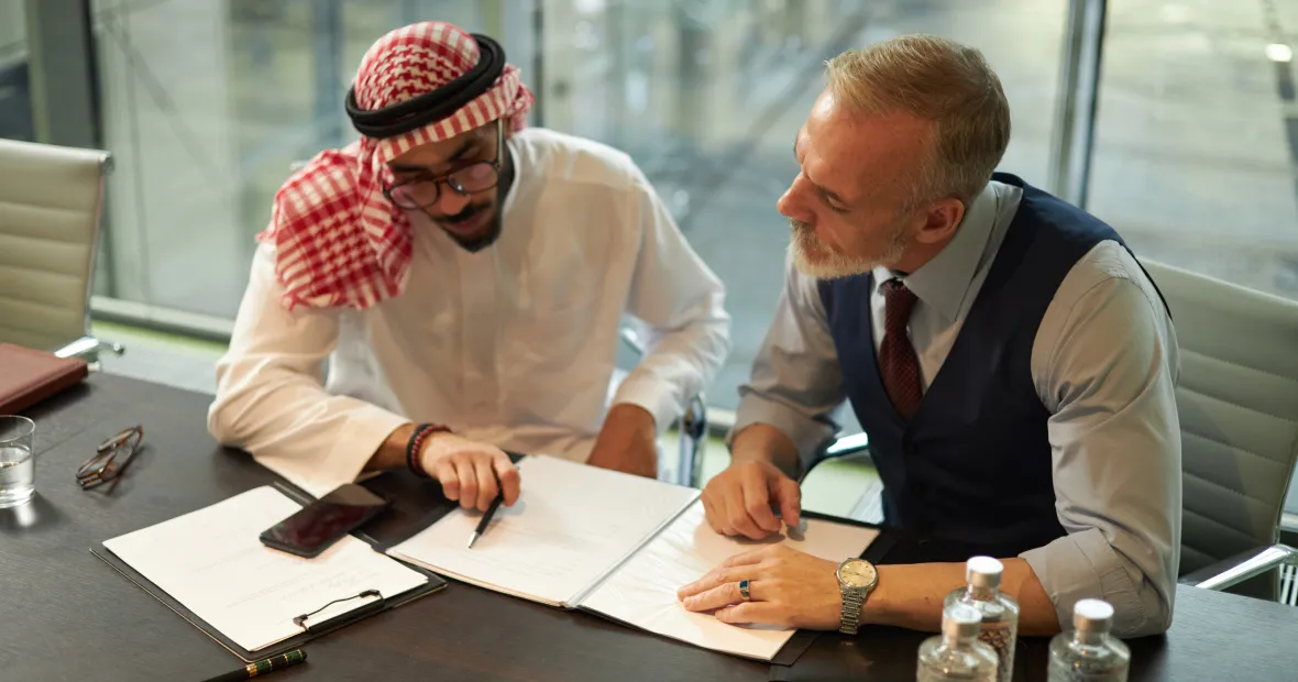 Two men sitting at a conference table reviewing documents together; one man wears traditional Middle Eastern attire and the other wears a dress shirt with vest and tie.