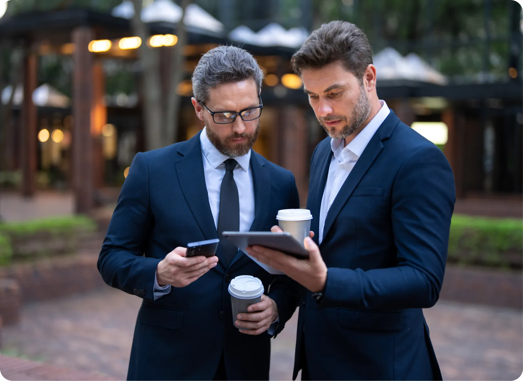Two businessmen in dark suits standing outdoors, each holding a coffee cup while looking at a smartphone and a tablet respectively.