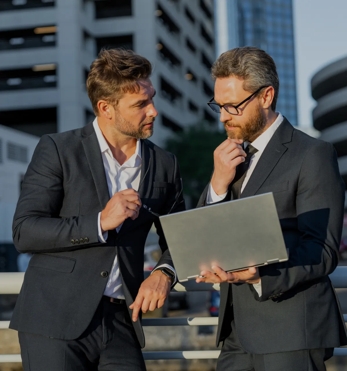 Two men in dark business suits stand outdoors in an urban setting, one holding a laptop and the other gesturing as they engage in a focused discussion.