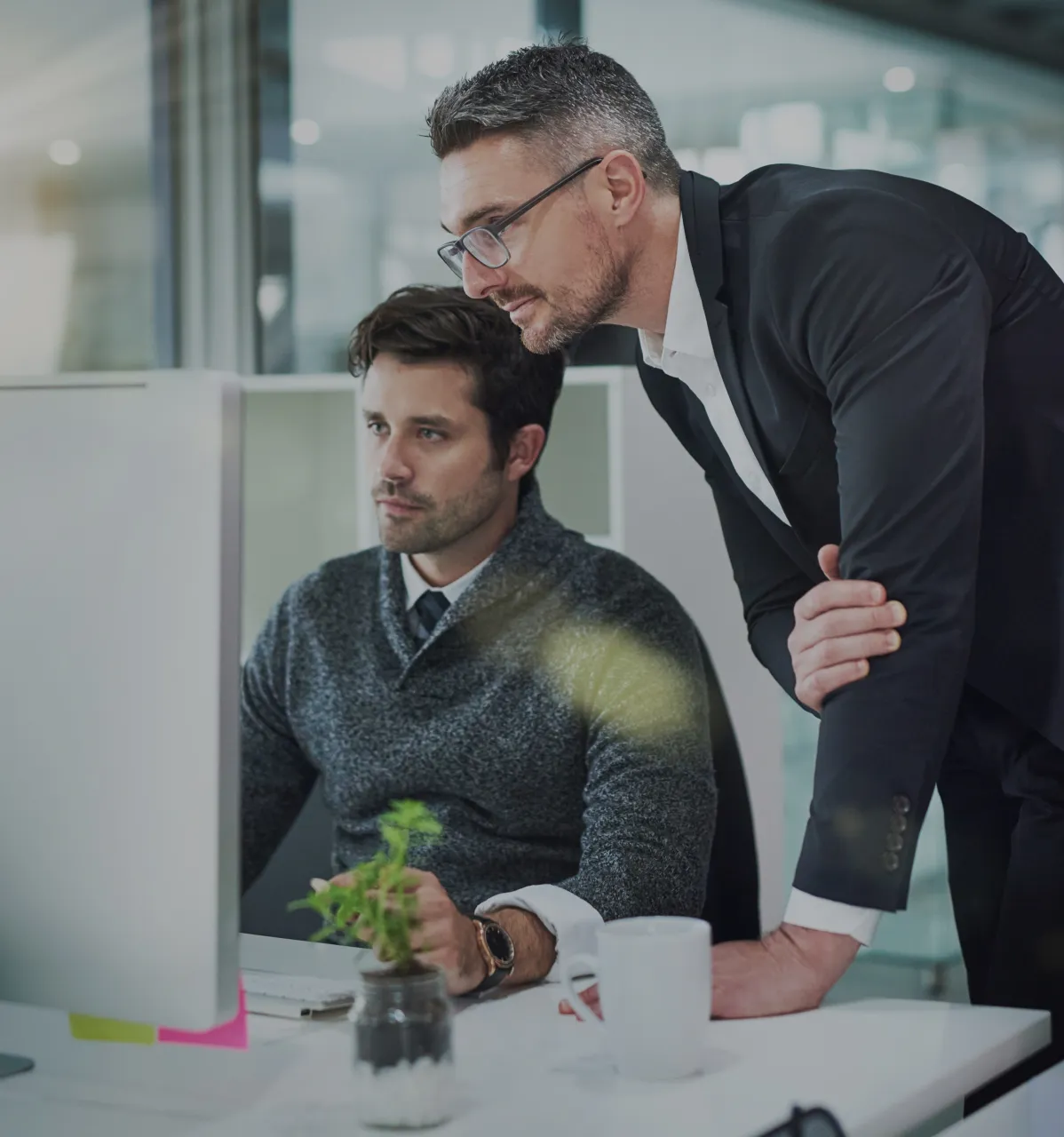 Two men in an office focused on a computer screen, one seated wearing a gray sweater and the other standing in a black suit and glasses leaning in to look at the monitor, with a small potted plant and a white mug on the desk.