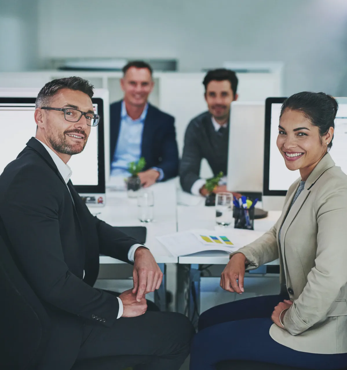 Four business professionals sitting at a white office desk with computer monitors, two in the foreground smiling towards the camera and two blurred in the background, in a modern office environment.