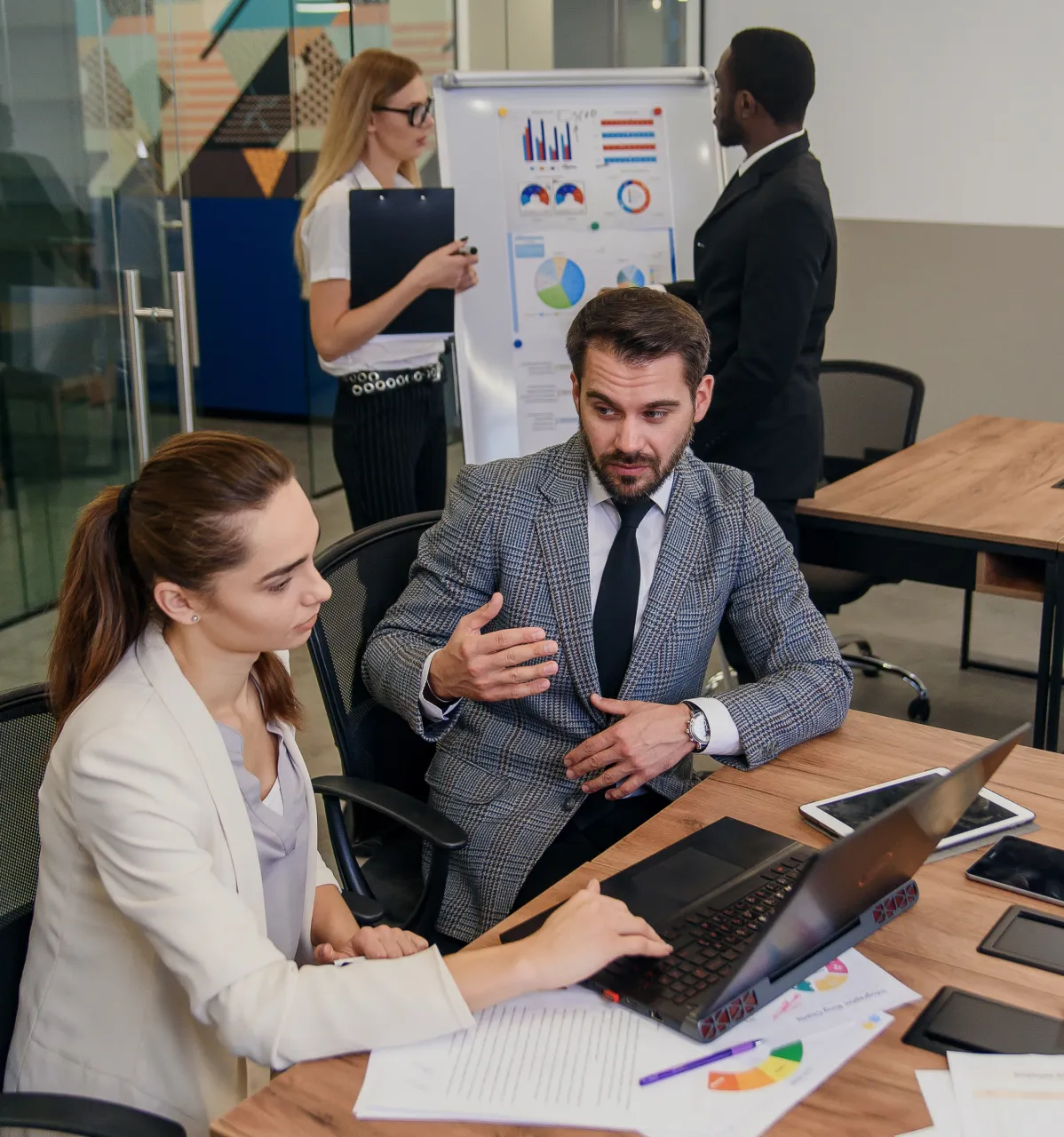 Two men and two women in a modern office collaborating; one woman and a man sit at a desk with a laptop and charts while another woman holds a clipboard and a man stands looking at a flipchart with graphs and pie charts.