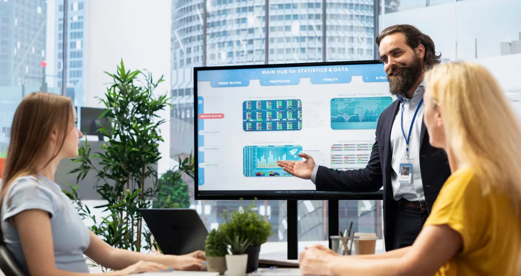 A man in business attire with a beard presents data charts on a large screen to two seated women in a modern office with large windows and plants.
