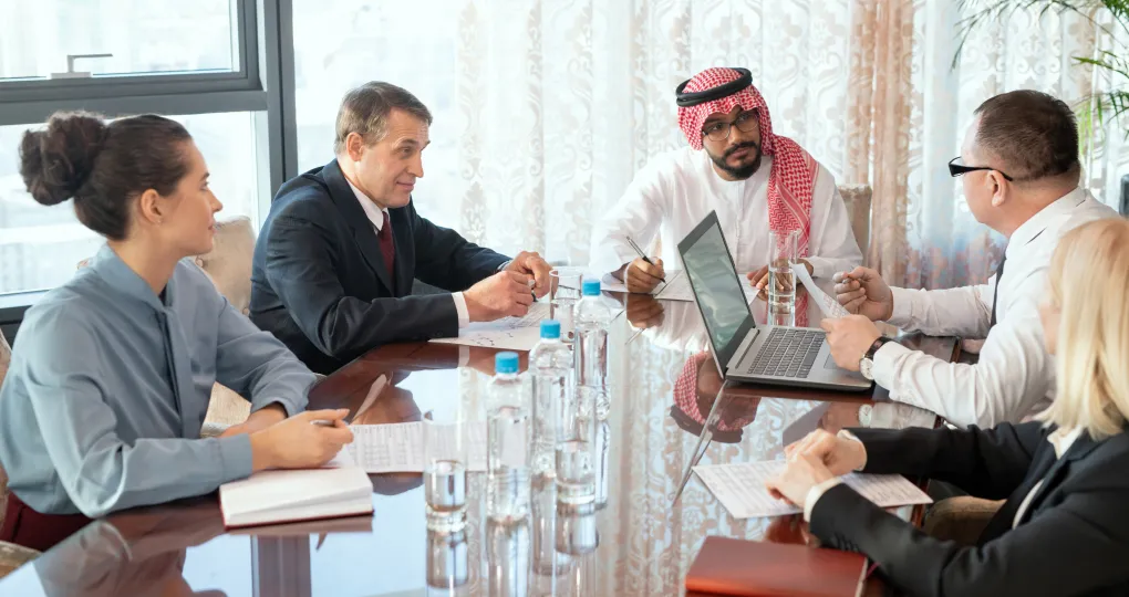 Five diverse business professionals engaged in a discussion around a conference table with laptops, notebooks, and water bottles, including a man in traditional Middle Eastern attire.